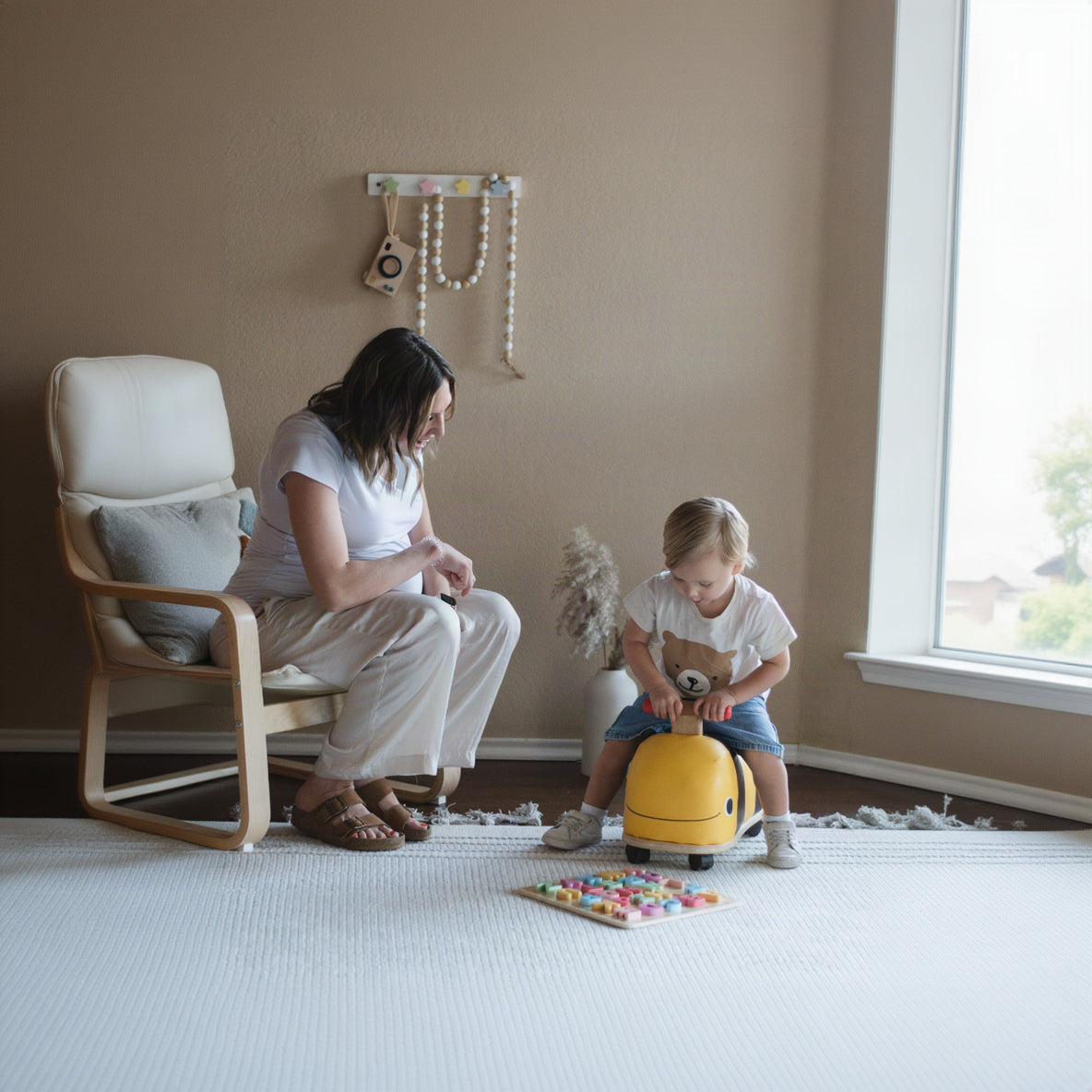 A woman sits on a wooden chair next to a young child riding a yellow toy suitcase in a bright, neutral room. The child is wearing a white t-shirt and blue jeans, and the woman is wearing a white t-shirt and light beige pants.
